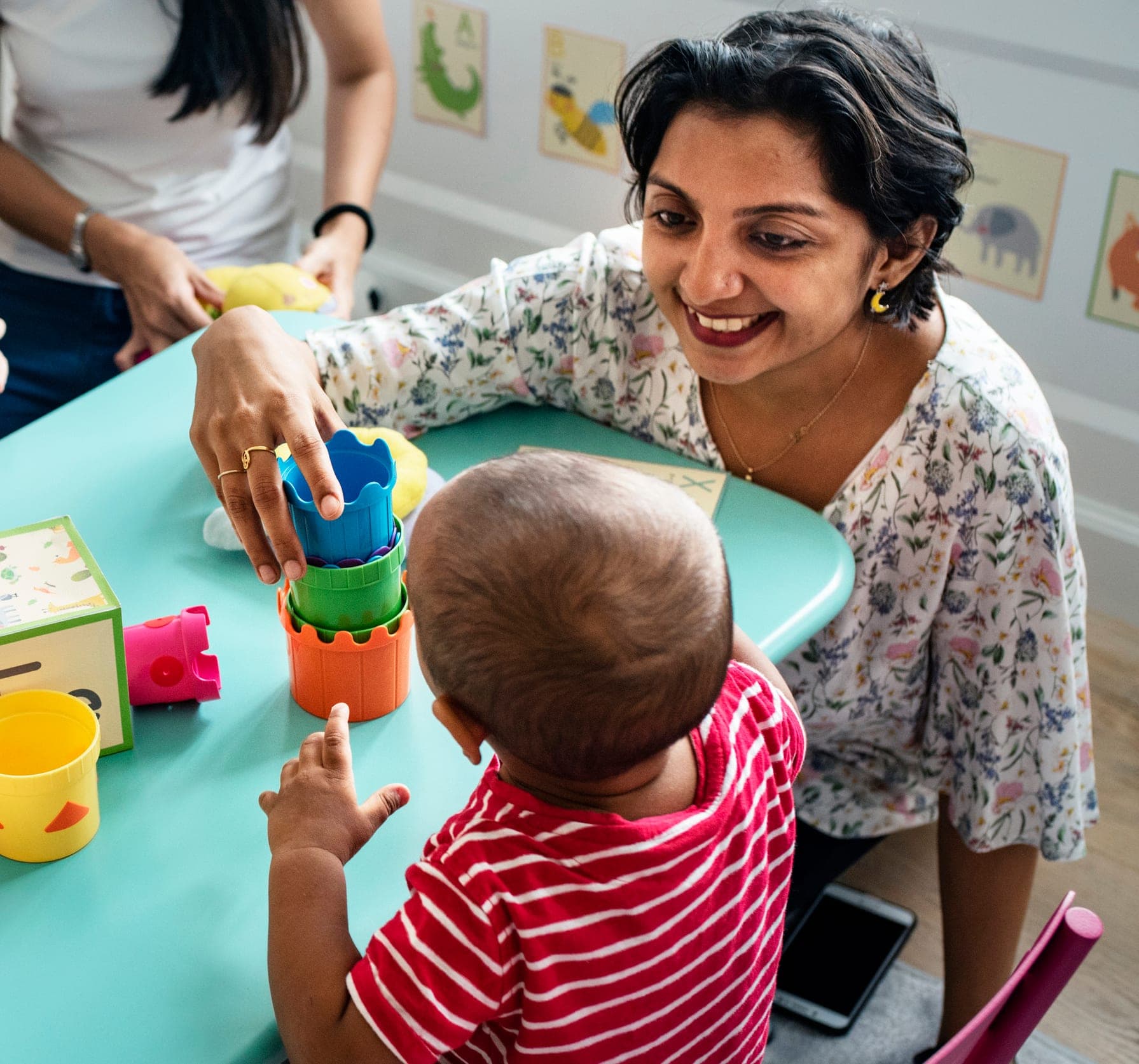 A teacher smiling with a toddler stacking colorful cups at a classroom table